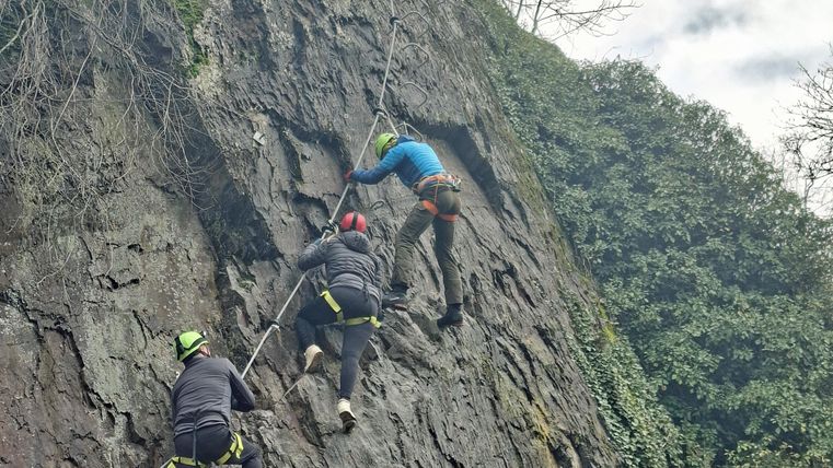 Drei Bergsteiger klettern an einer steilen Felswand. Der Himmel ist leicht bewölkt und Bäume umgeben die Szene.