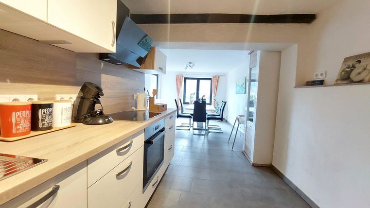 A modern kitchen with light cabinets and wooden shelves. In the background, there is a dining area with a table and chairs.