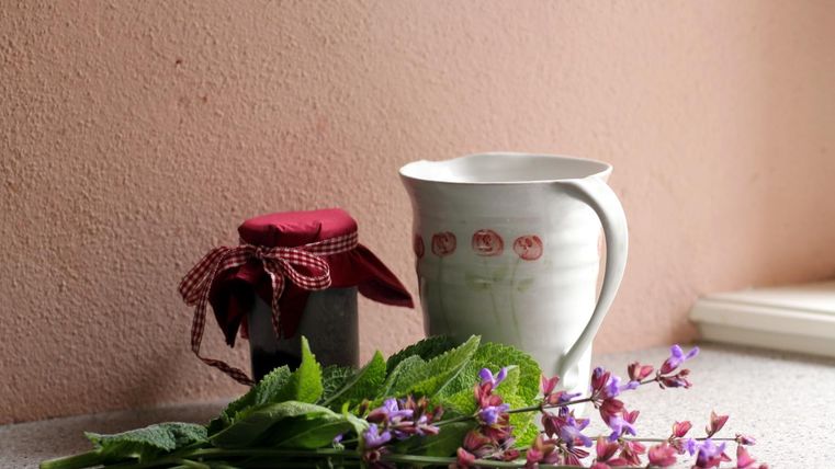 Ein hübscher Keramikkrug steht neben einem Glas mit rotem Deckel. Daneben liegen frische Kräuter und bunte Blumen.