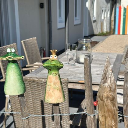 Two colorful figures in the shape of frogs hanging on a fence. In the background, a cozy terrace with garden furniture can be seen.