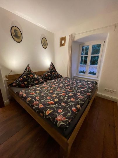 A cozy bedroom with a double bed and colorful cushions. Large windows provide a view outside and there are decorative wall pictures.