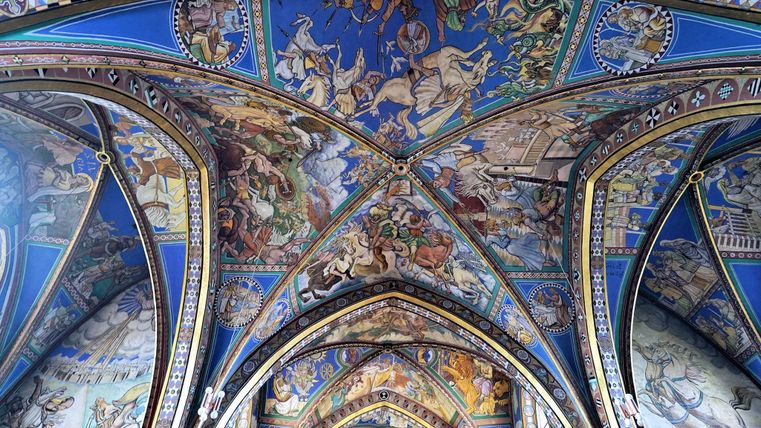 Eine kunstvolle Decke mit bunten Fresken in einem gotischen Gebäude. Die Bilder zeigen historische Szenen und Figuren in lebendigen Farben.