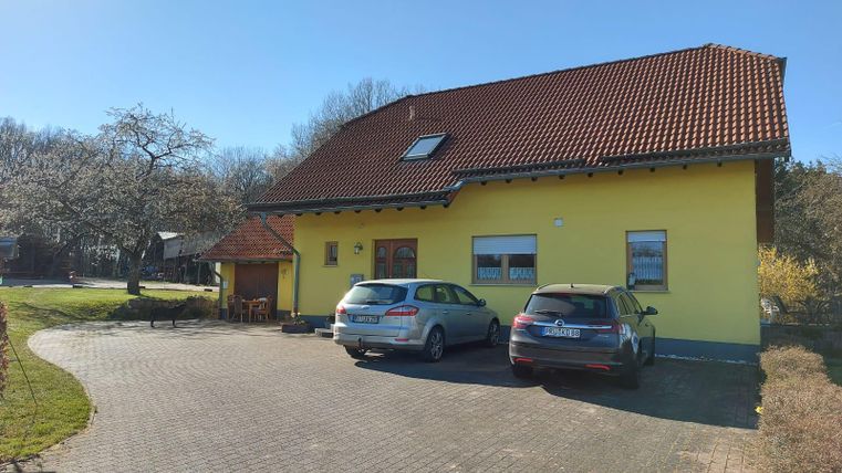 Een geel huis met een rood dak staat aan een geplaveide oprit. Twee auto's staan geparkeerd voor het gebouw, omgeven door groene natuur.