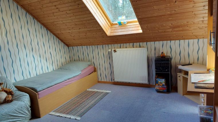Een gezellige zolderkamer met een eenpersoonsbed en een bureau. De kamer heeft een schuine wand en een raam dat veel licht binnenlaat.