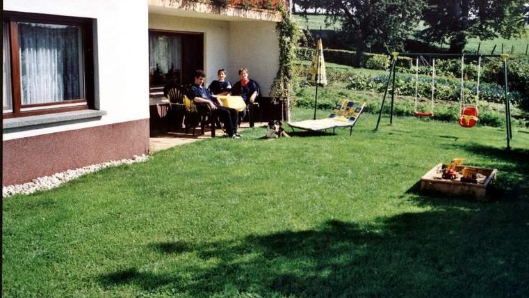Eine grüne Rasenfläche hinter einem Haus mit einer Terrasse. Drei Personen sitzen am Tisch und entspannen sich, während sich im Hintergrund eine Schaukel und ein kleiner Garten befinden.