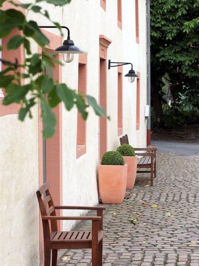 Eine gemütliche Straße mit alten Steinplatten und einer hellen Wand. Zwei Holzbänke und Töpfe mit grünen Pflanzen sorgen für eine einladende Atmosphäre.