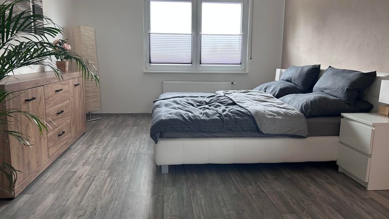 Ein modernes Schlafzimmer mit einem großen Bett und dunkler Bettwäsche. Eine Kommode und ein Pflanze ergänzen die Einrichtung.