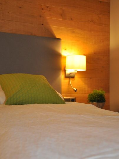 Ein gemütliches Schlafzimmer mit einem Betthaupt und einer grünen Decke. An der Wand hängt eine Lampe und eine kleine Pflanze steht auf dem Nachttisch.