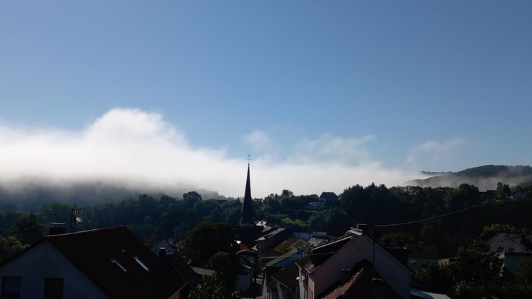 Eine malerische Aussicht mit Häusern im Vordergrund und einem Kirchturm. Im Hintergrund ist dichter Nebel über den Hügeln und ein blauer Himmel zu sehen.