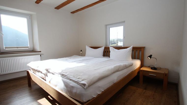 Ein helles Schlafzimmer mit einem großen Doppelbett und zwei Fenstern. Die Wände sind in einem schlichten Weiß gehalten, und es gibt Holzbalken an der Decke.