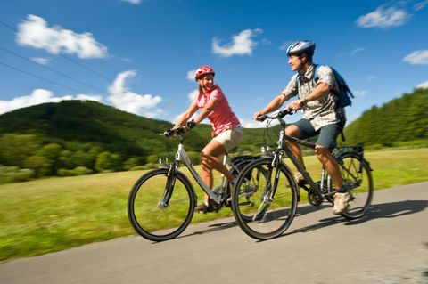 Ein Paar fährt zusammen mit Fahrrädern auf einem Weg durch eine grüne Landschaft. Der Himmel ist blau mit ein paar Wolken.