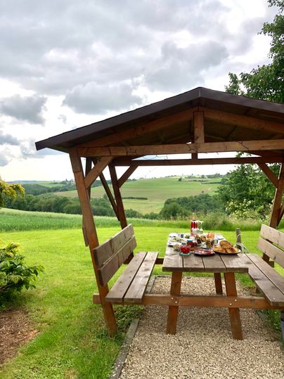Un pavillon confortable avec une table de pique-nique sur une prairie verte. À l'arrière-plan, des collines douces s'étendent sous un ciel nuageux.