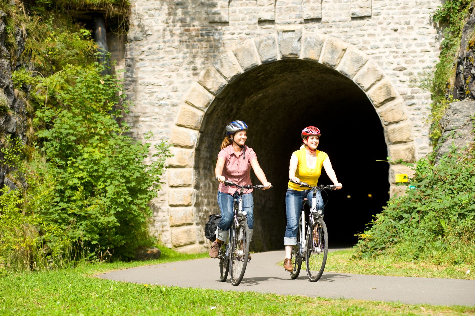 Zwei Frauen fahren mit ihren Fahrrädern auf einem Weg, der durch eine Steintunnelöffnung führt. Umgeben von üppigem Grün und sonnigem Wetter wirkt die Szene einladend und freundlich.