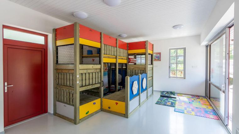 Ein heller Spielraum mit bunten Spielgerüsten und einem leicht zugänglichen Eingang. Der Raum ist gut beleuchtet und kindgerecht gestaltet.