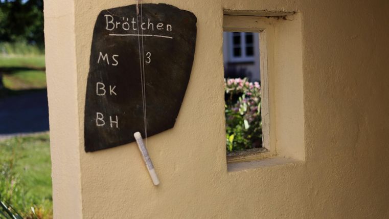 Eine Tafel mit der Aufschrift „Brötchen“ und verschiedenen Abkürzungen ist an einer Wand befestigt. Neben der Tafel befindet sich ein kleines Fenster.