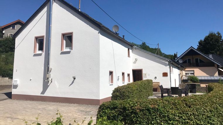 Ein modernes, weißes Haus mit einem kleinen Garten und Terrasse. Im Hintergrund sind weitere Wohngebäude und ein klarer blauer Himmel zu sehen.