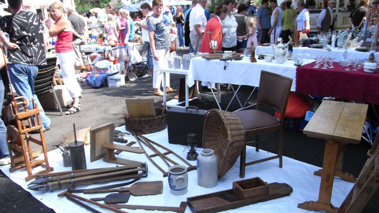 Ein belebter Flohmarkt mit vielen Ständen und Besuchern. Verschiedene alte Möbel und Haushaltsgegenstände sind auf einer weißen Decke ausgestellt.