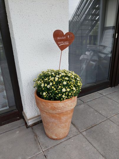A blooming flower pot with yellow and white flowers stands in front of a window. A heart-shaped sign with the inscription "Welcome" adorns the pot.