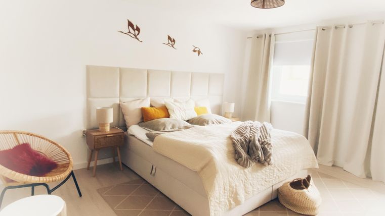 Een lichte slaapkamer met een groot bed en decoratieve kussens. Er is een gezellige stoel en veel daglicht door het raam.