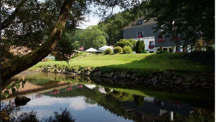Eine ruhige Landschaft mit einem klaren Fluss, der von Bäumen umgeben ist. Im Hintergrund sieht man ein Gebäude und grüne Wiesen.