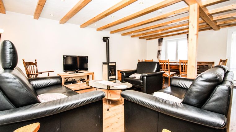 Ein gemütliches Wohnzimmer mit zwei schwarzen Ledersofas und einem Couchtisch. Ein Fernseher und ein Esstisch sind ebenfalls vorhanden, sowie sichtbare Holzdeckenbalken.