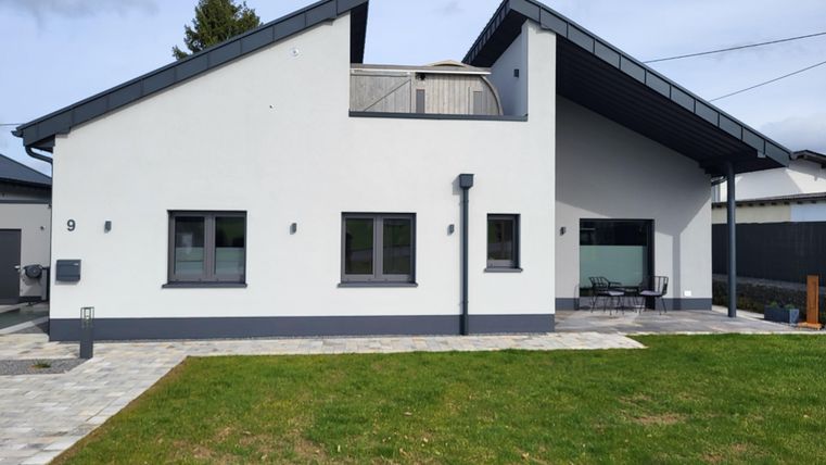 Ein modernes, einstöckiges Haus mit einem grauen Dach und großen Fenstern. Der Garten ist grün und gepflegt, mit einem gepflasterten Bereich vor dem Haus.