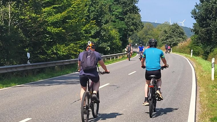 Zwei Radfahrer fahren auf einer ruhigen Landstraße entlang. Im Hintergrund sind weitere Radfahrer und grüne Bäume zu sehen.