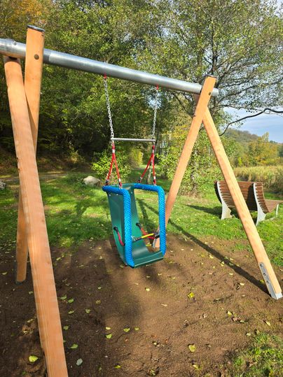 Een kleurrijke schommel staat in een groen speelgebied. Het speelplein is omringd door bomen en heeft een rustige sfeer.