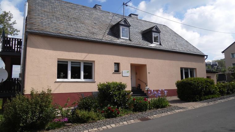 Een twee verdiepingen tellend, muurkleurig huis met een grijs dak. Het heeft grote ramen en is omringd door verzorgde planten.