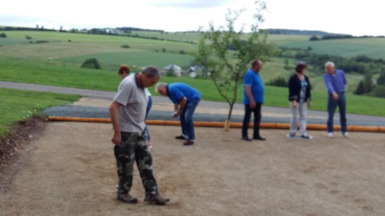 Un groupe de personnes joue à la pétanque sur une place en gravier en plein air. À l'arrière-plan, on peut voir un paysage verdoyant avec des collines.