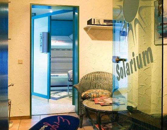 Een ontvangstgedeelte van een zonnestudio met een gezellige stoel en een tafel. Achter een glazen deur is het zonnebank apparaat zichtbaar.