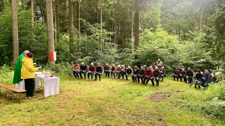 Een groep mensen zit in het bos en luistert naar een presentatie. Voor hen staat een tafel met materialen en een persoon spreekt.