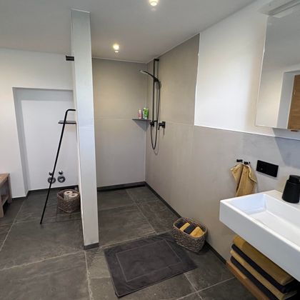 A modern bathroom with a gray tiled floor and white walls. There is a shower, a sink, and a small wooden table.