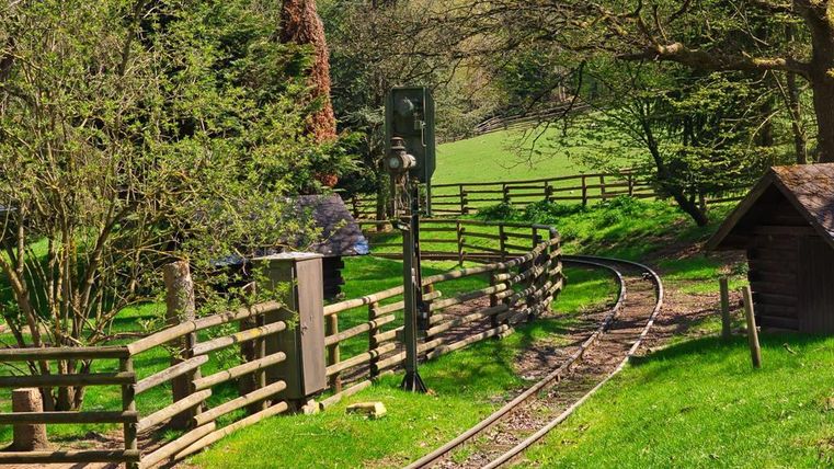Ein malerischer Weg mit einer kleinen Eisenbahnschiene, umgeben von grünen Wiesen und Bäumen. Im Hintergrund ist ein Signal und ein Holzhaus sichtbar.