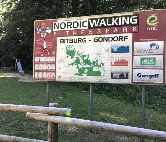 Sign for the Bitburg-Gondorf Nordic Walking Fitness Park with map and sponsor logos in the forest, next to a playground with slide., © TI Bitburger Land