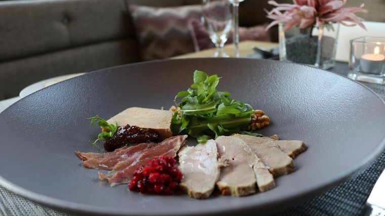 Ein elegantes Gericht auf einem flachen Teller, bestehend aus verschiedenen Fleischsorten, frischem Salat und einer Beilage aus roten Beeren. Im Hintergrund sind stilvolle Tischdekoration und Glaswaren zu sehen.
