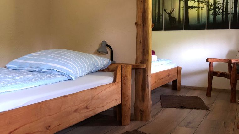 Une chambre simple avec deux cadres de lit en bois et une petite table. Un tableau d'une forêt est accroché au mur.