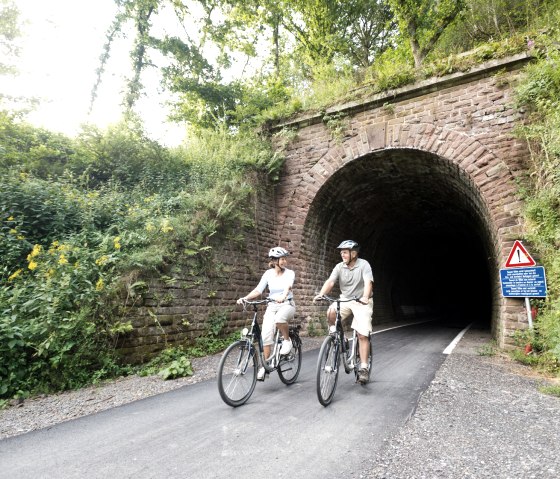 Twee fietsers rijden uit een overwoekerde tunnel van de Vennbahn bij Lommersweiler. Er staat een waarschuwingsbord aan de rand., © vennbahn.eu
