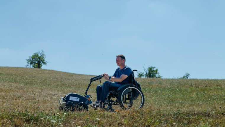 Ein Mann sitzt in einem Rollstuhl auf einer blühenden Wiese. Im Hintergrund ist ein blauer Himmel und einige Bäume zu sehen.