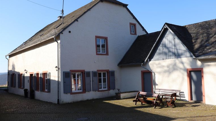 Ein weißes Haus mit grauen Fensterläden und roten Akzenten. Im Vordergrund steht eine Holzbank mit Tisch.