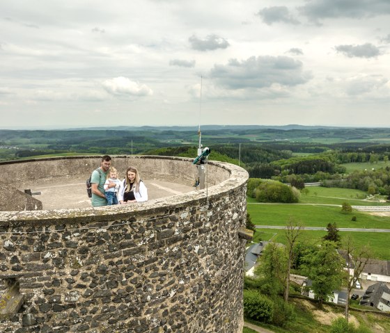 Eine Familie steht auf einem Turm in der Eifel, umgeben von grüner Landschaft und bewölktem Himmel.
