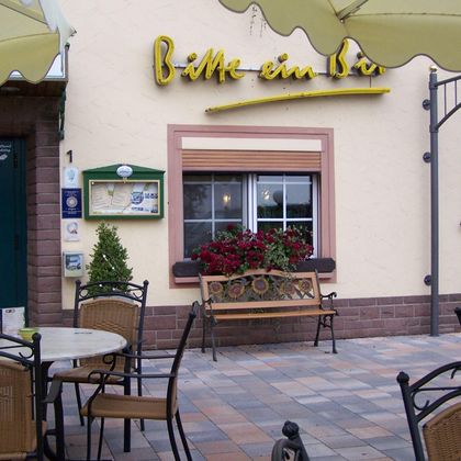 Eine gemütliche Außenterrasse mit Tischen und Stühlen vor einem kleinen Restaurant. Das Schild des Restaurants zeigt den Namen „Bitte ein Bier“ und blühende Pflanzen schmücken das Fenster.