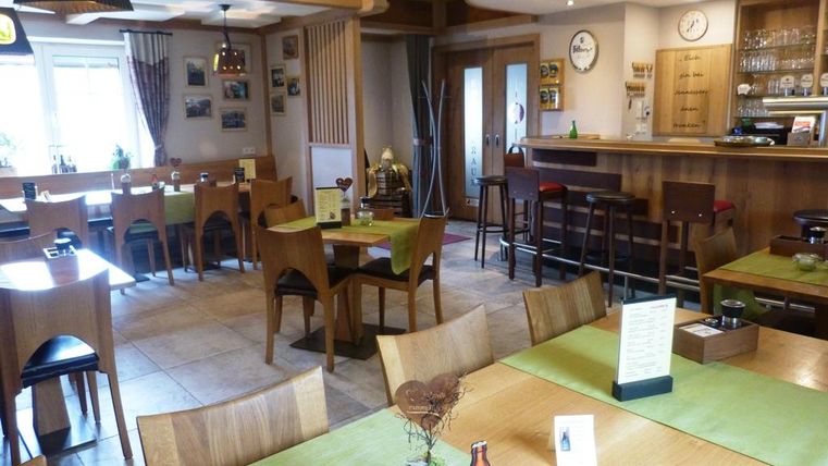 Ein gemütliches Restaurant mit Holzmöbeln und grünen Tischdecken. An den Wänden hängen Bilder und eine Bar ist im Hintergrund sichtbar.