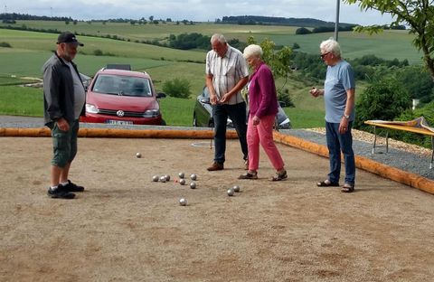 Un groupe de quatre personnes joue à la pétanque sur un terrain de sable. En arrière-plan, on peut voir un paysage verdoyant et une voiture rouge.