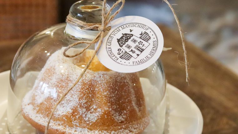 Ein frisch gebackener Kuchen unter einer Glasglocke, dekoriert mit Puderzucker. Eine Etikette hängt am Glas mit Informationen zum Produkt.