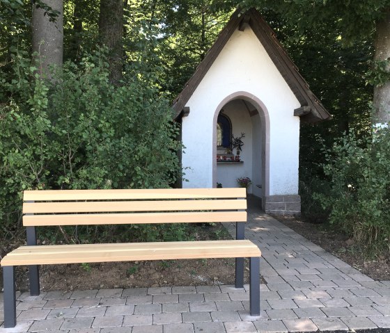A small white chapel with a brown roof stands at the edge of the forest. In front of it is a wooden bench on a paved path., © TI Bitburger Land
