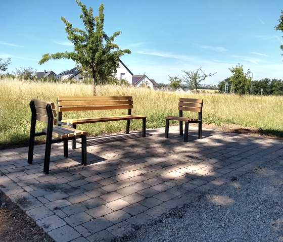 Three wooden benches stand on a paved square in a rural setting. Trees, a meadow and houses can be seen in the background., © TI Bitburger Land