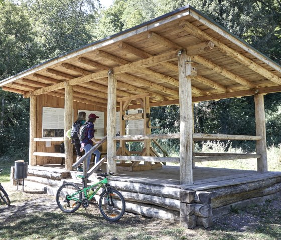 Zwei Personen lesen Infotafeln in einem hölzernen Unterstand im Grünen. Fahrräder stehen daneben, umgeben von Bäumen., © TI Hocheifel-Nürburgring_Jonathan_Andrews