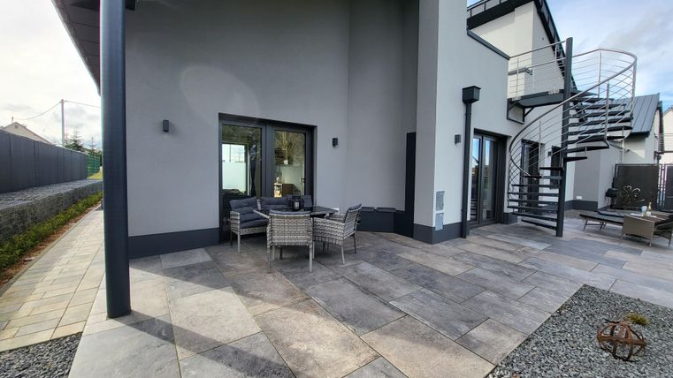 Eine moderne Terrasse mit bequemen Möbeln und einem Blick auf ein zweigeschossiges Gebäude. Die Umgebung ist gepflegt mit Steinplatten und Kies.