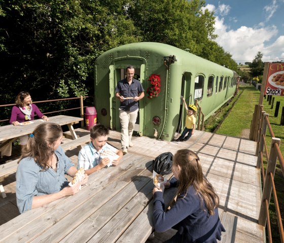 Familien genießen Waffeln an Holztischen neben einem grünen Eisenbahnwaggon in einer ländlichen Umgebung., © vennbahn.eu